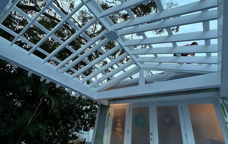 White pergola beams framing a view of trees above a building with glass doors at dusk.