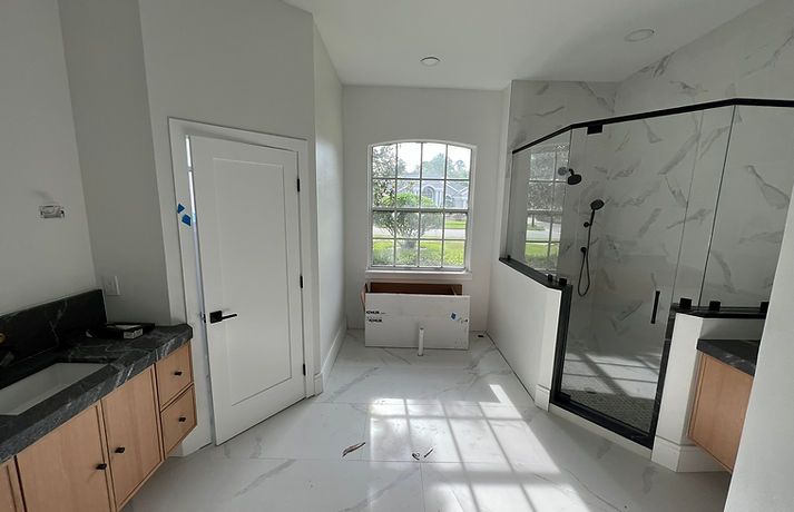 Modern bathroom with light wood vanities, marble-look floor tiles, a glass shower stall, and a central window.