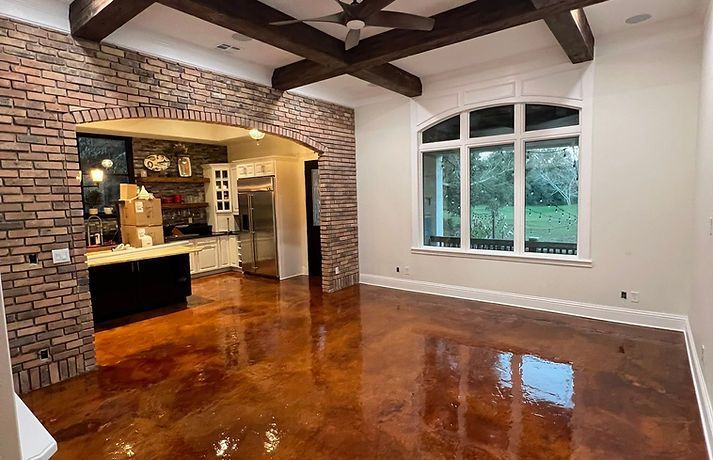 Living room with brown stained concrete floors, vaulted wood-beam ceiling, and an arched brick entryway to the kitchen.