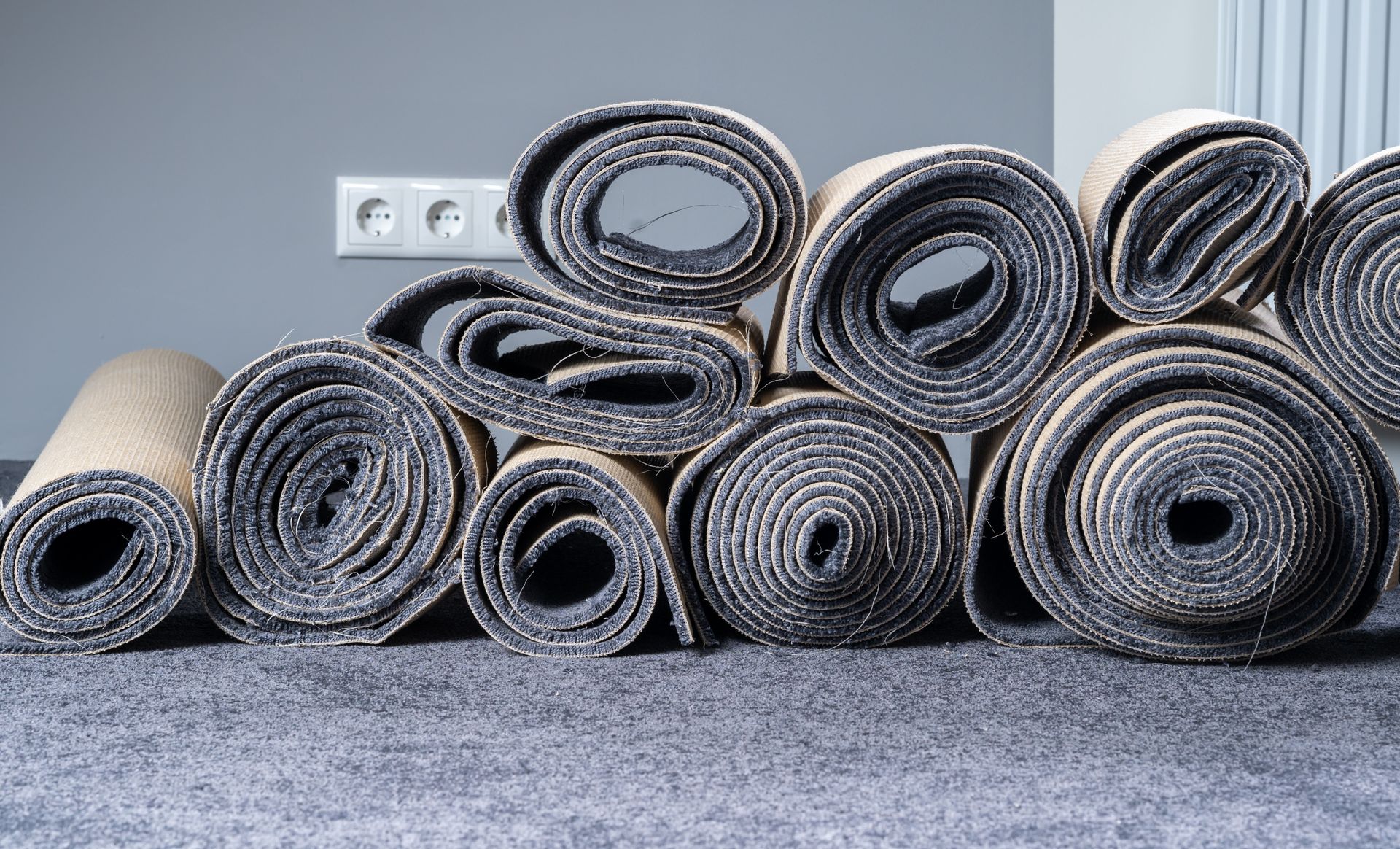 Rolled-up carpets of various sizes stacked against a gray wall with an electrical outlet.