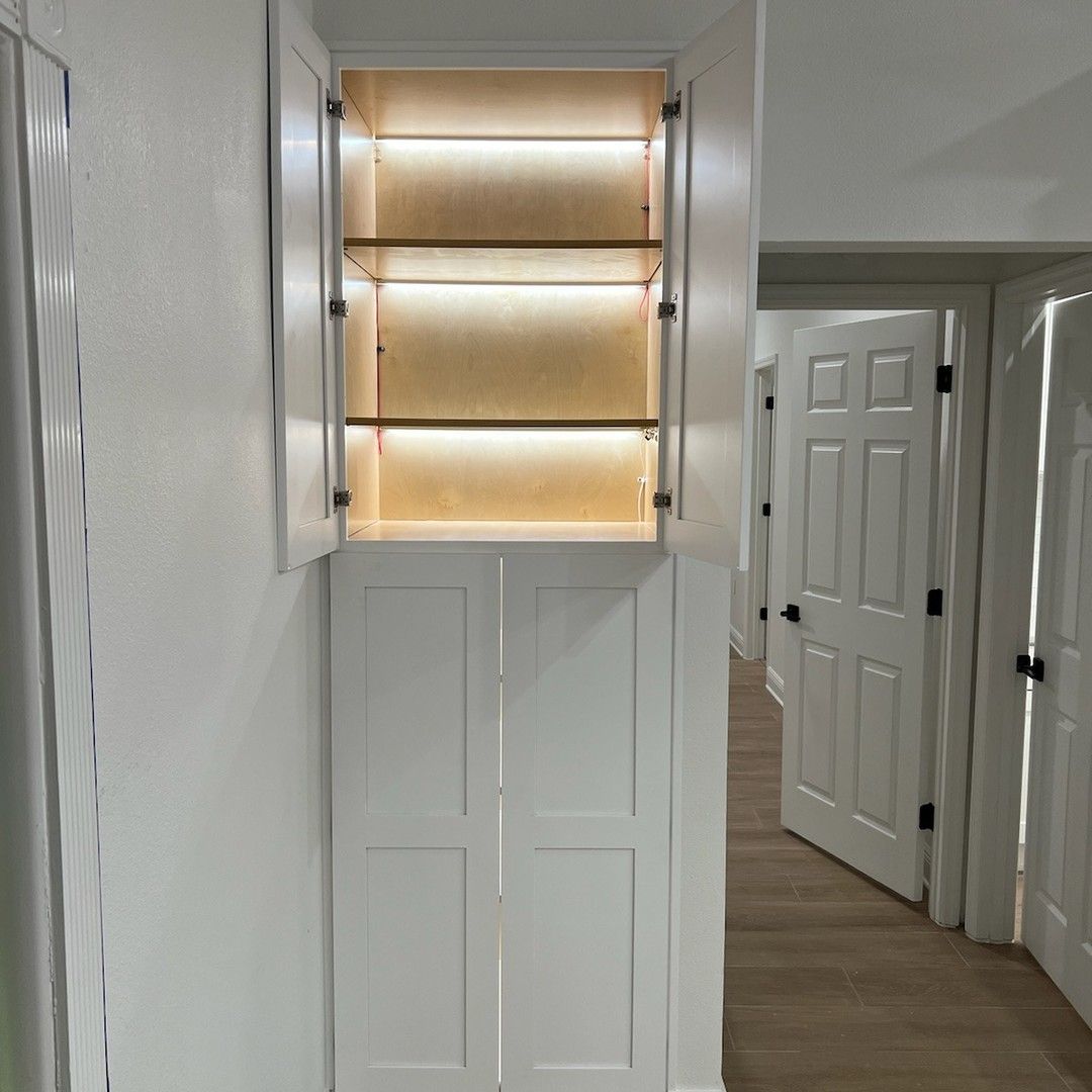 White cabinet with open upper doors and interior lighting, two glass shelves.  Lower cabinet doors are closed.  Hallway in the background.