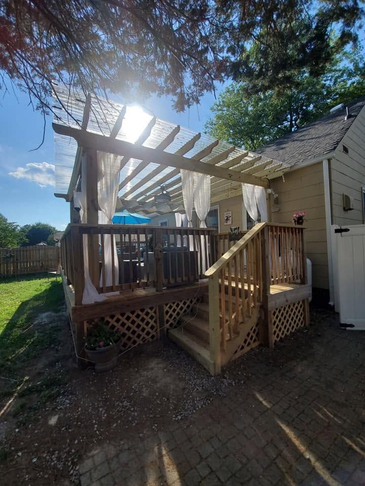 Backyard deck with pergola, stairs, and railing beside a beige house in bright sunlight