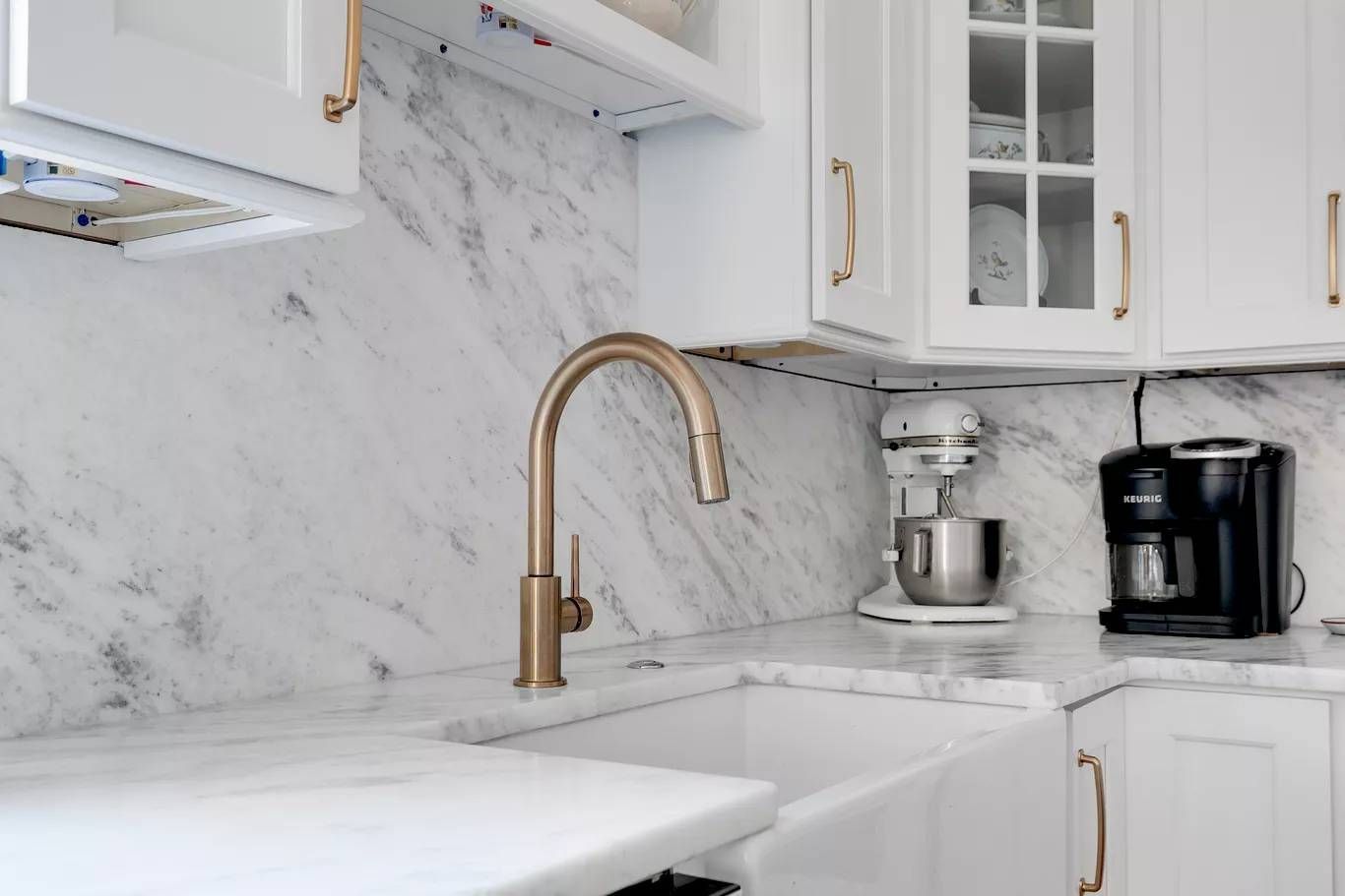 White kitchen with marble backsplash, brass faucet, and black coffee maker on the counter