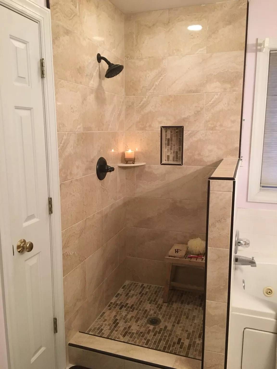 Small tiled shower with a built-in shelf, wall-mounted showerhead, candle, and patterned floor mat.