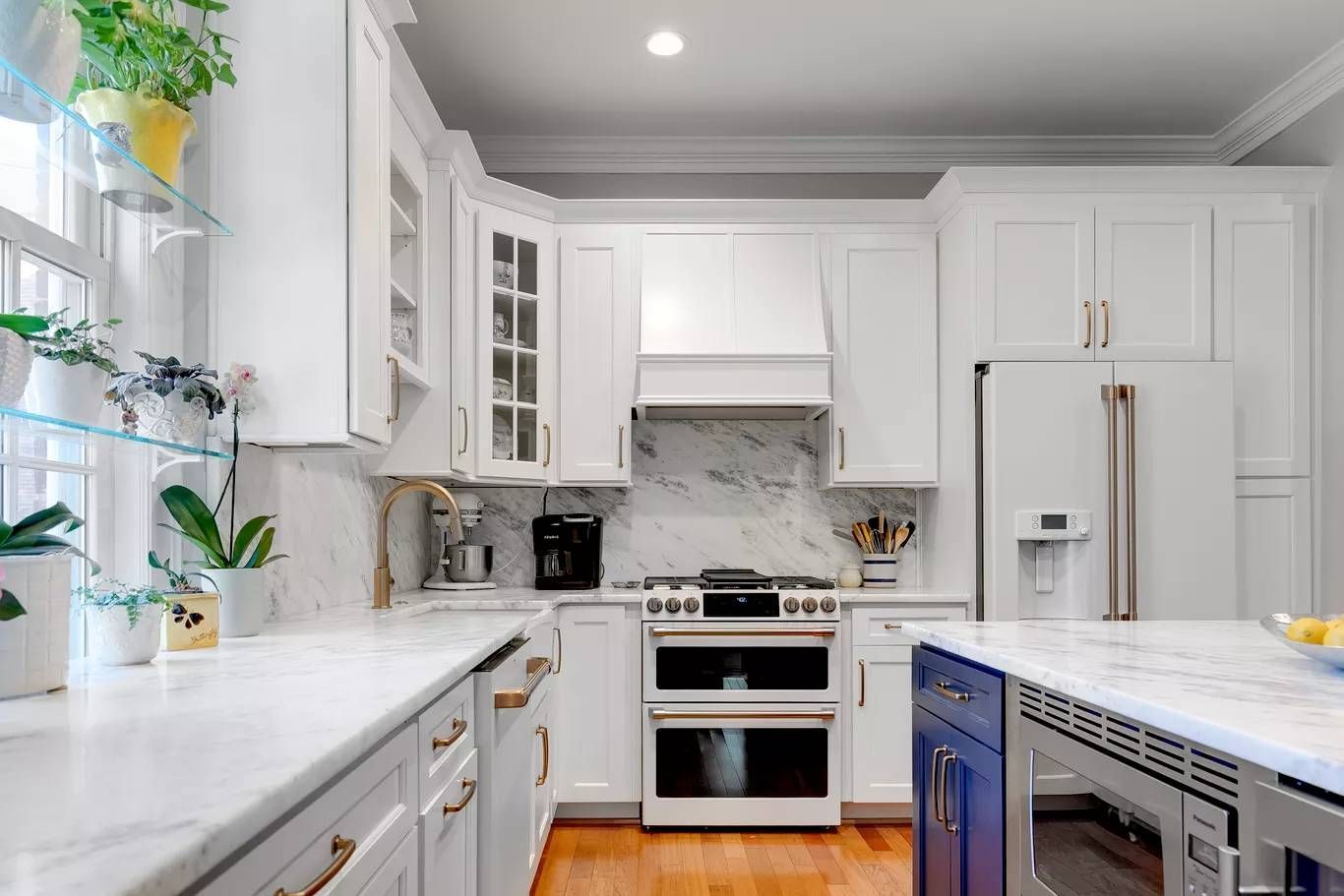 Bright white kitchen with marble counters, blue island, stainless steel appliances, and potted plants by the window