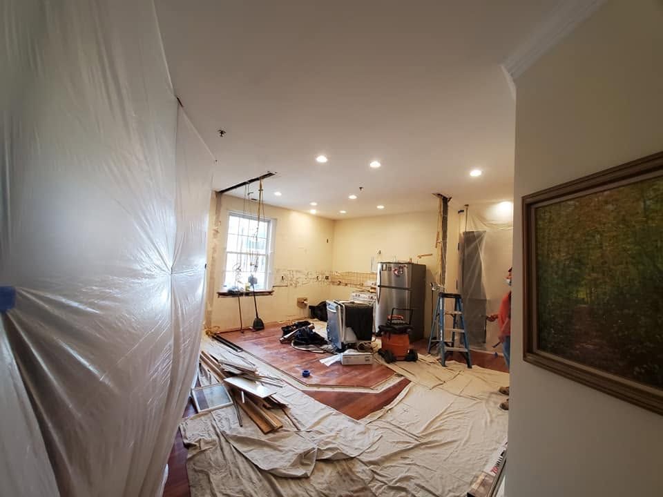 Room under renovation with plastic sheeting, tools, rugs, and a ladder in a bright living space.