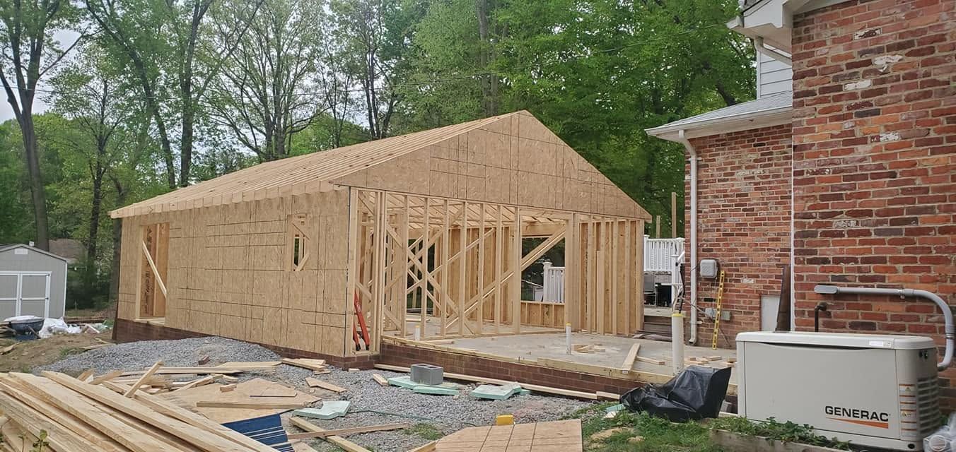 House under construction with framed walls and roof sheathing beside a brick building, wood and debris in foreground