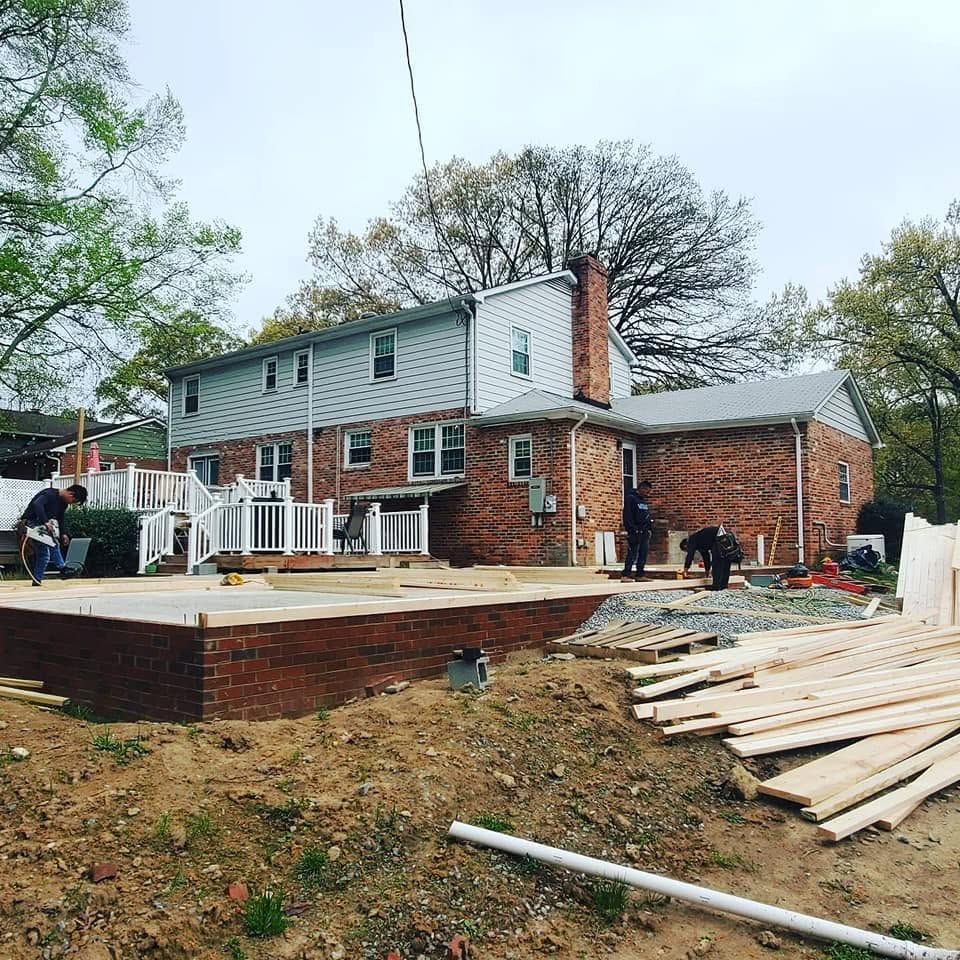 House exterior under renovation with workers, new deck framing, and lumber stacked in the foreground