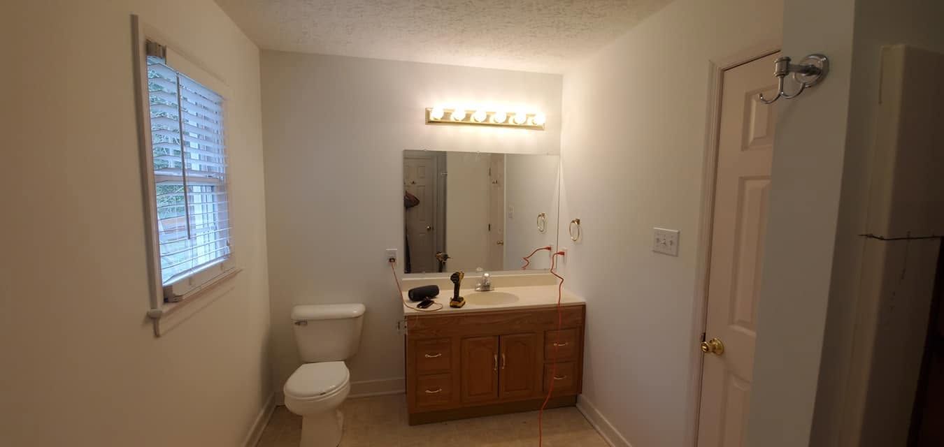 Small bathroom with toilet, wood vanity and mirror, window on the left, and a door on the right.