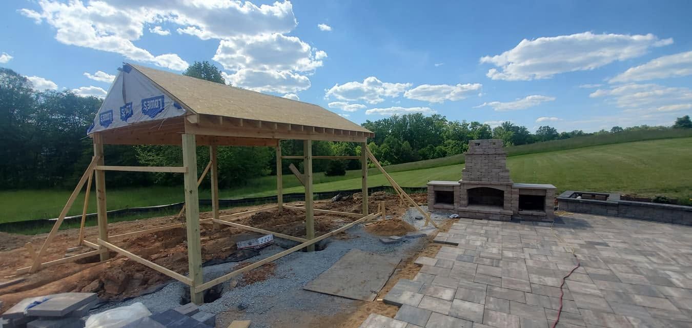 Partially built wooden pavilion beside a stone fireplace on a concrete slab in a grassy field