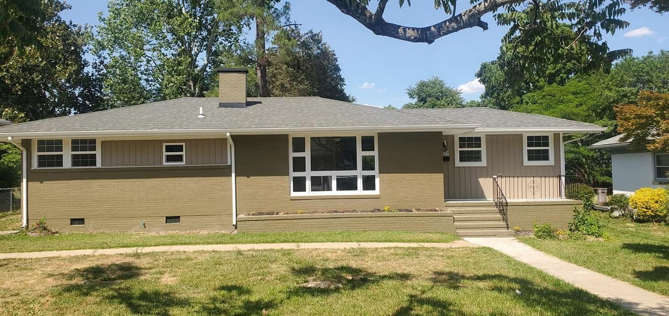 Single-story tan brick house with a front walkway, lawn, and large tree in a sunny yard