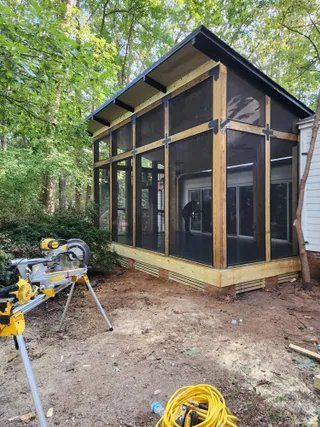 Partially built screened porch with black framing, glass doors, and construction tools in a wooded yard