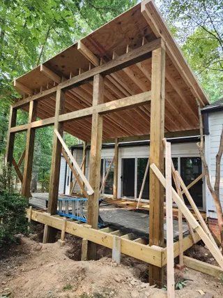 Wooden deck frame under construction beside a house, with tall posts, beams, and diagonal braces.