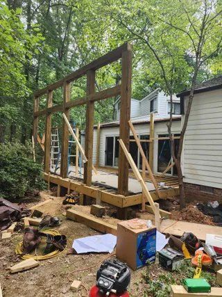 Wooden wall frame under construction beside a house in a wooded yard, with tools and lumber on the ground.