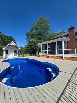 Blue in-ground pool on a wooden deck beside a brick house under a clear sky