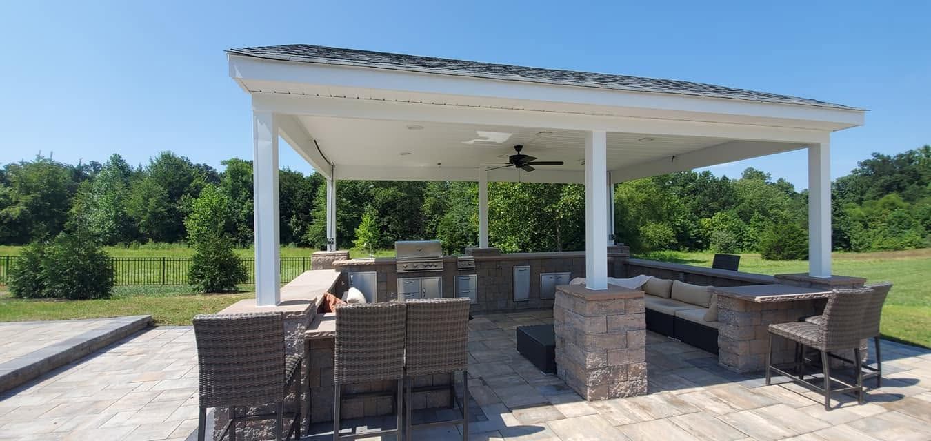 Outdoor pavilion with stone bar, wicker chairs, and picnic tables on a sunny patio by greenery