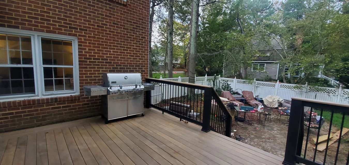 Wooden deck beside a brick house, with a stainless grill and black railing overlooking a wooded yard.