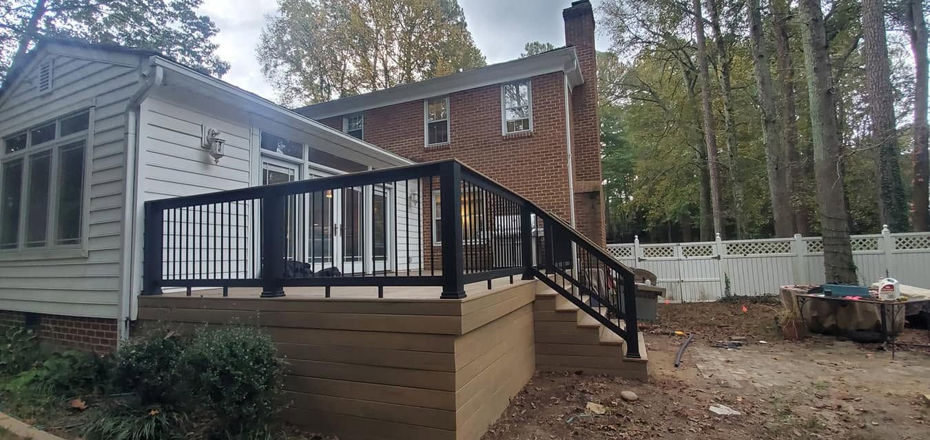 Small house deck with black railing and stairs beside a white siding addition in a wooded yard