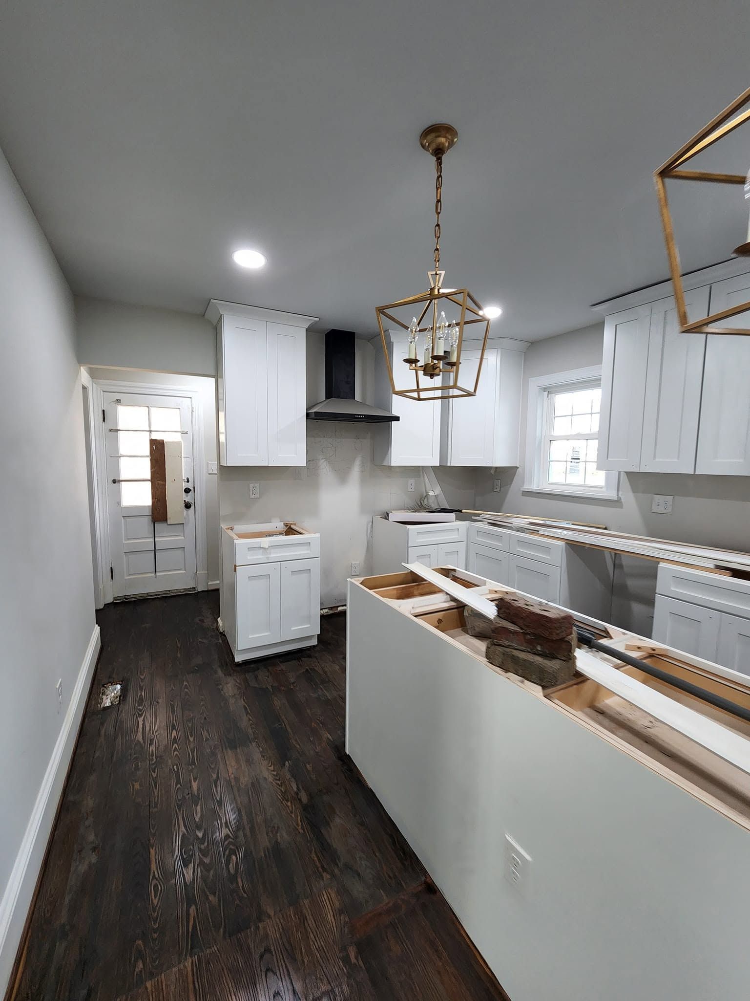 Bright white kitchen with dark wood floors, gold chandelier, island, cabinets, and hallway view