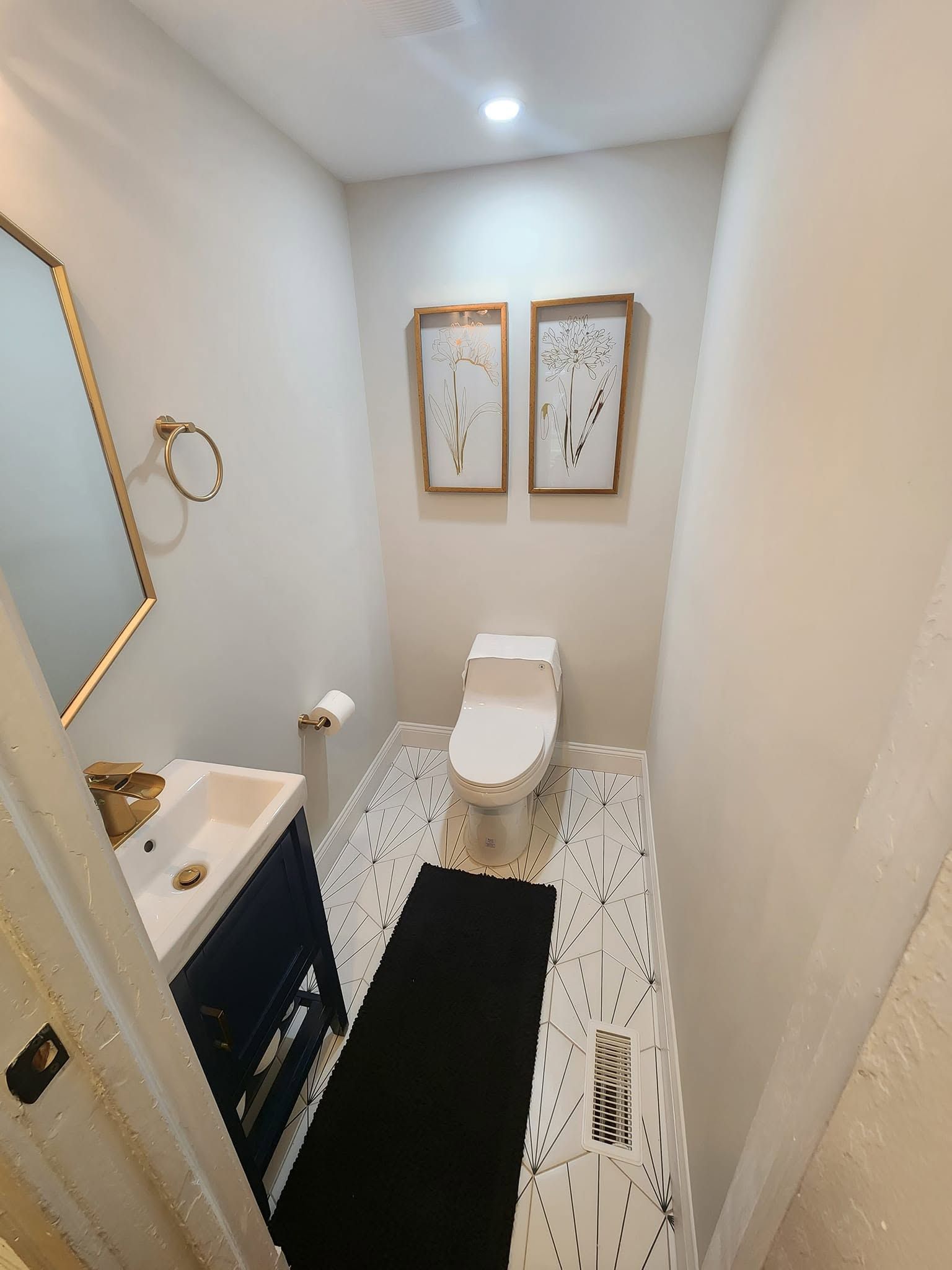 Small narrow bathroom with white walls, patterned floor, black runner rug, sink, toilet, and framed wall art.