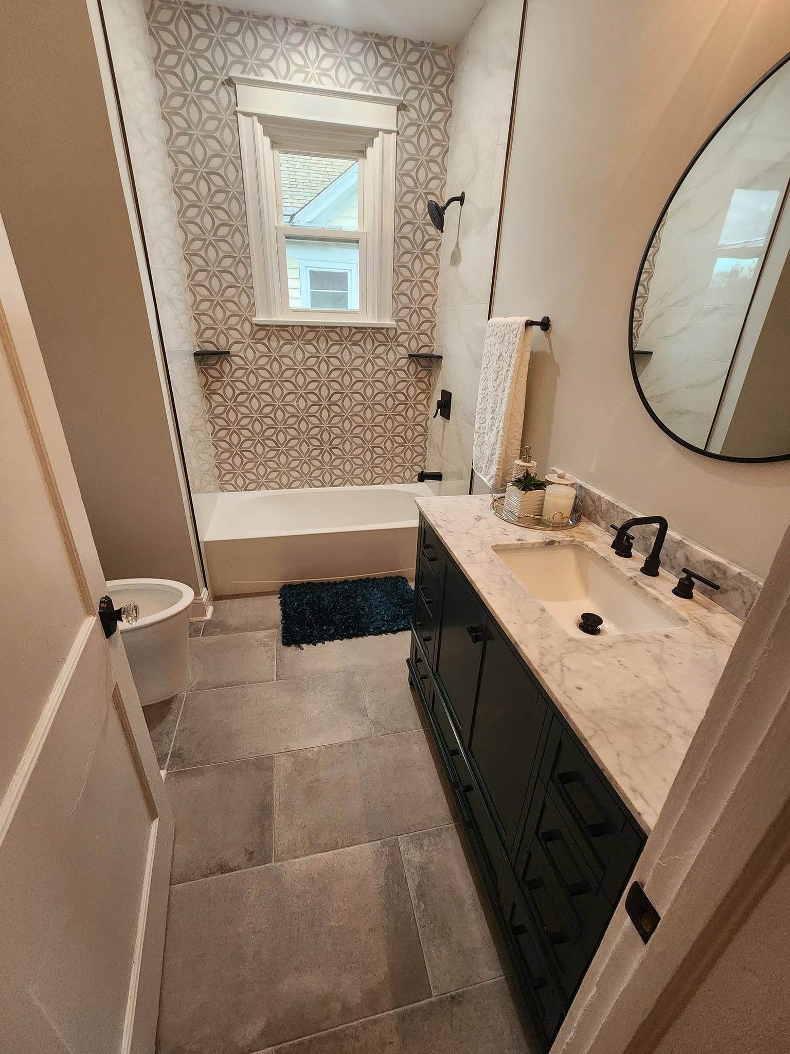 Narrow bathroom with tiled floor, vanity sink, round mirror, tub, and window at the far end.