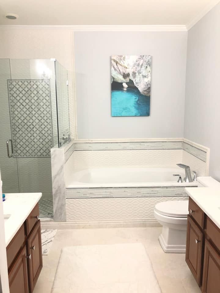 Bright bathroom with white tile, wooden vanities, glass shower, and a tub beneath a wall print.