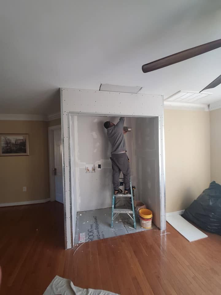 Person on a ladder painting a white wall opening in a living room with hardwood floors.