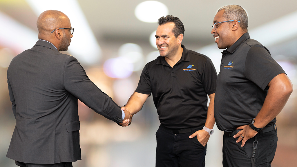 Three men in black attire shaking hands in a bright indoor setting, smiling and chatting.