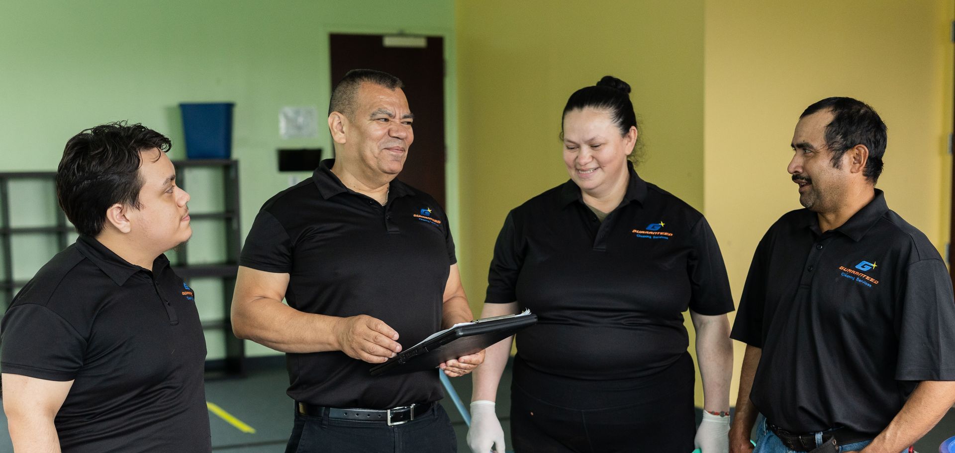 Four people in black shirts talking in a gym, one holding a tablet.
