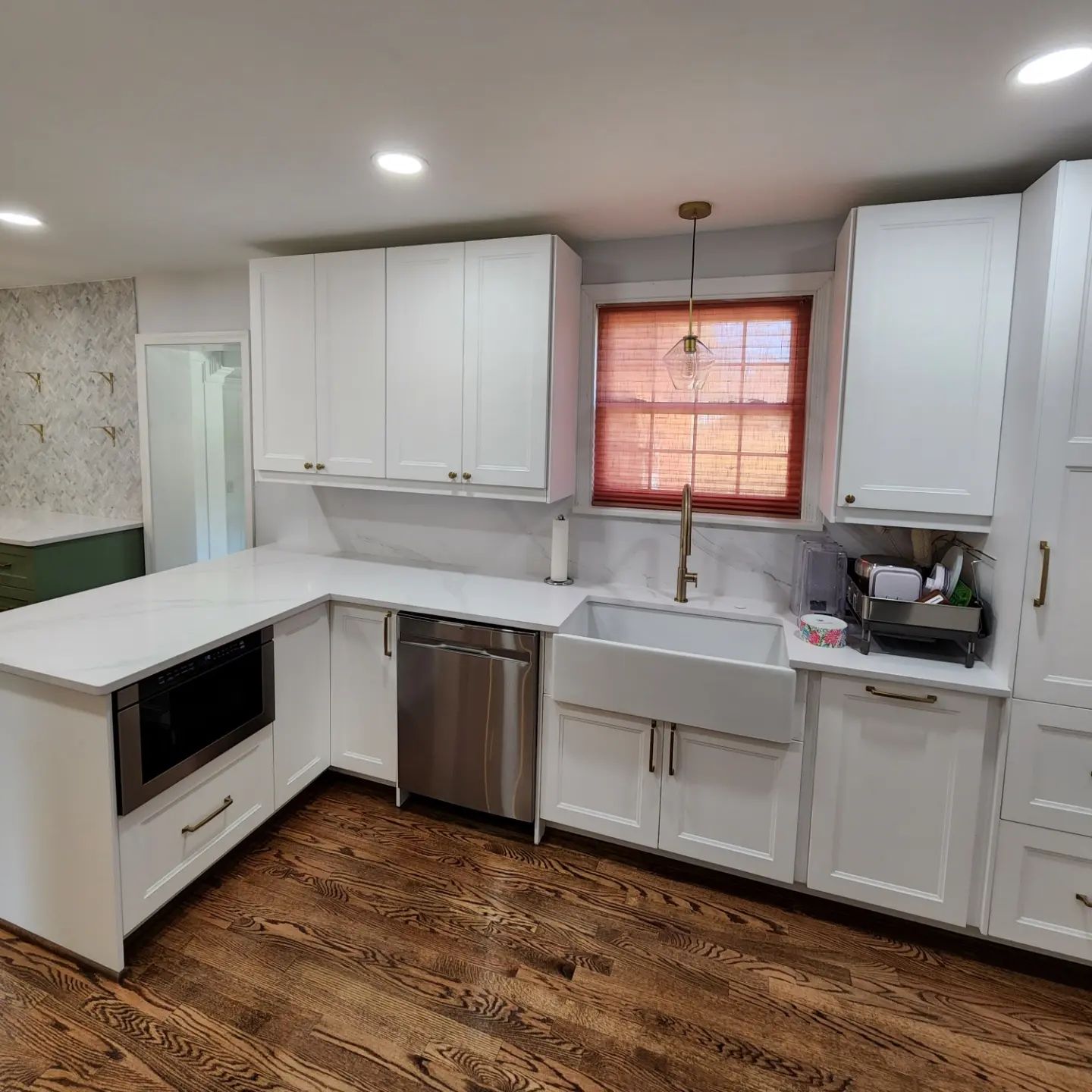 Bright white kitchen with L-shaped counters, farmhouse sink, wooden floor, and window with red blind.