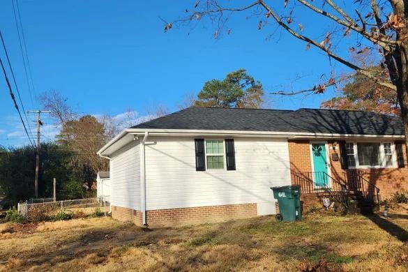 Single-story white house with a teal door and green trash bin in a sunny yard