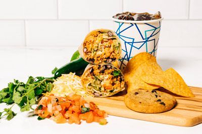 a burrito, tortilla chips, a cookie, and a cup of soda on a wooden cutting board