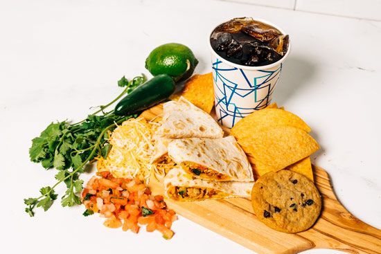 a wooden cutting board topped with quesadillas , tortilla chips , a cookie and a cup of soda .