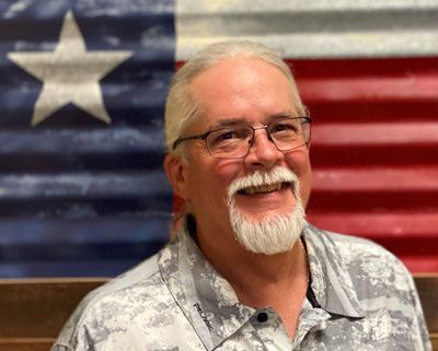 Man with glasses and white beard smiling, Texas flag backdrop.