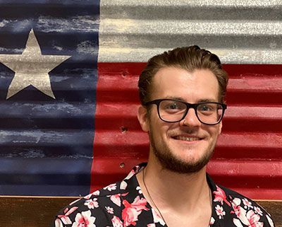 Man wearing glasses and floral shirt smiling in front of Texas flag.
