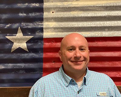 Man in blue plaid shirt smiling, standing in front of a Texas flag made of corrugated metal.