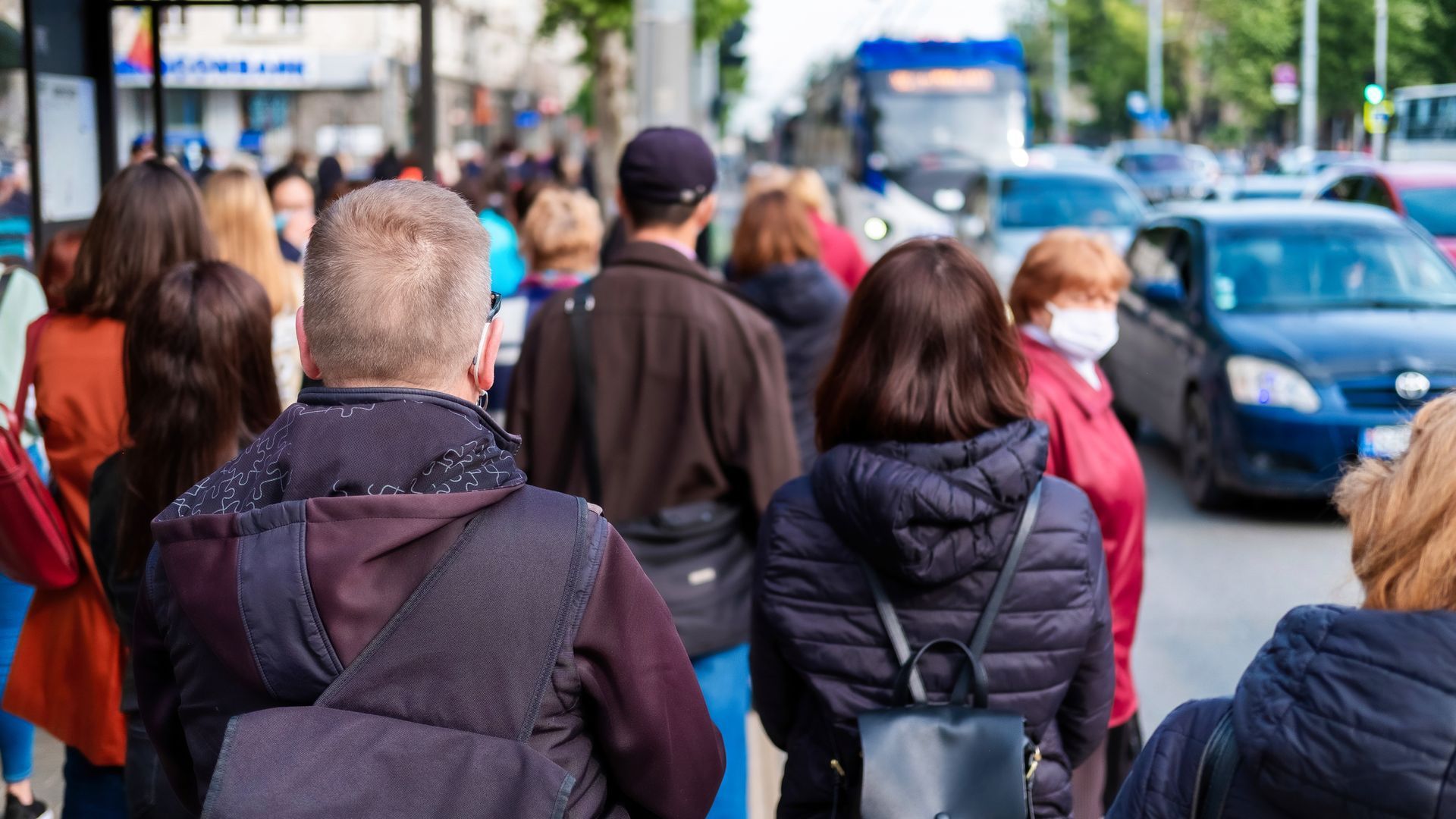 Folk går på et fortov i nærheden af trafik, hvor én person bærer en rød ansigtsmaske.