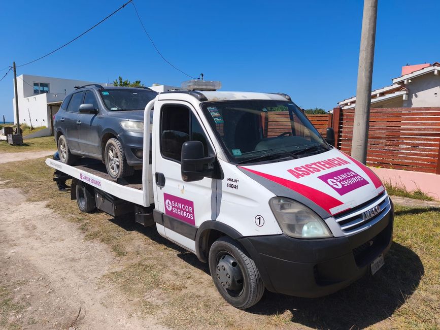 Una grúa roja transporta un SUV plateado en una plataforma por un camino rural de tierra bajo un cielo azul.