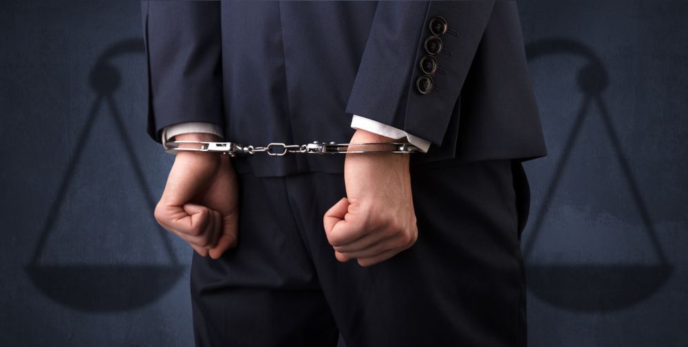 Man in suit with hands cuffed behind back, in front of scales of justice silhouette.