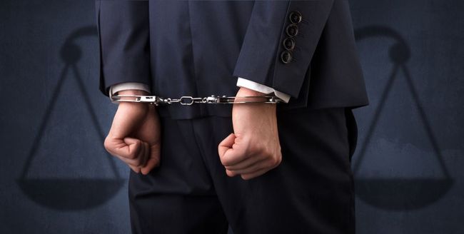 Man in suit with hands cuffed behind back, in front of scales of justice silhouette.