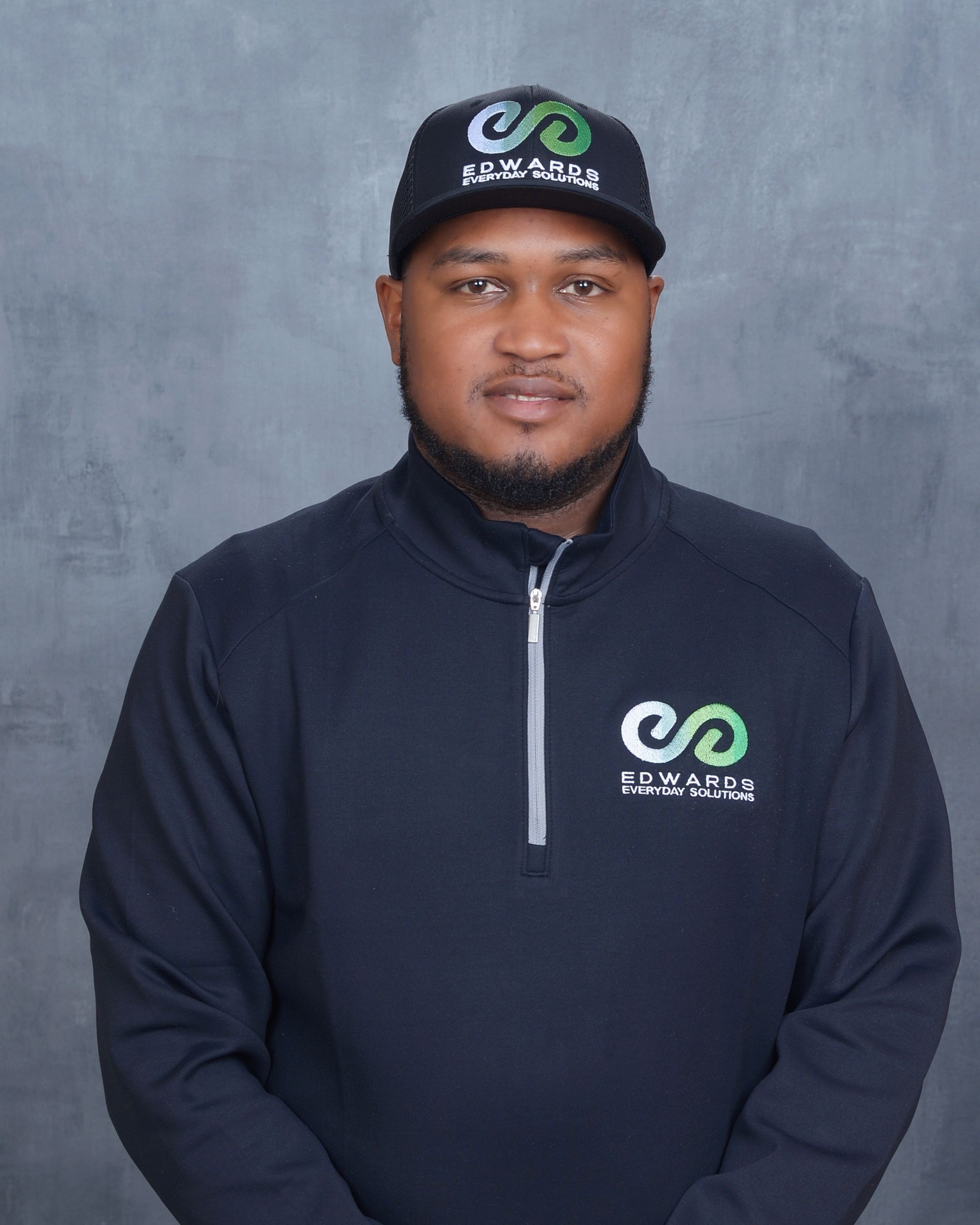 Man wearing a black cap and navy pullover with logo, standing against a gray background.