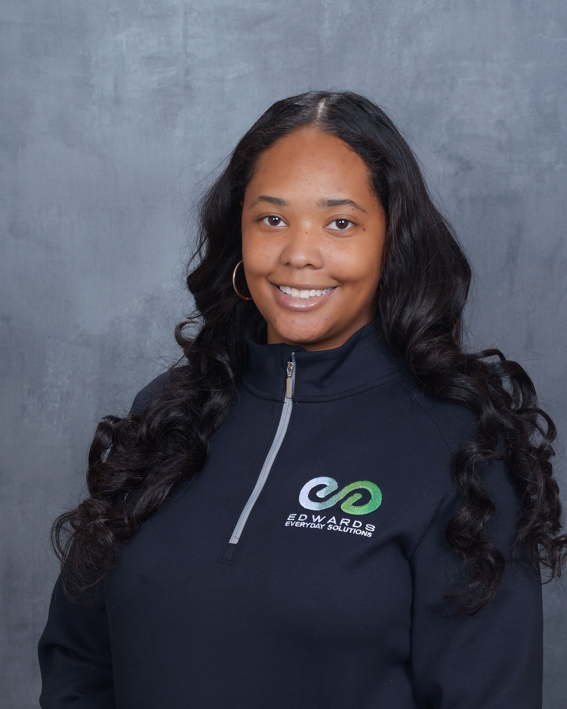 Smiling person in a black zip-up top with a logo, standing before a gray backdrop.