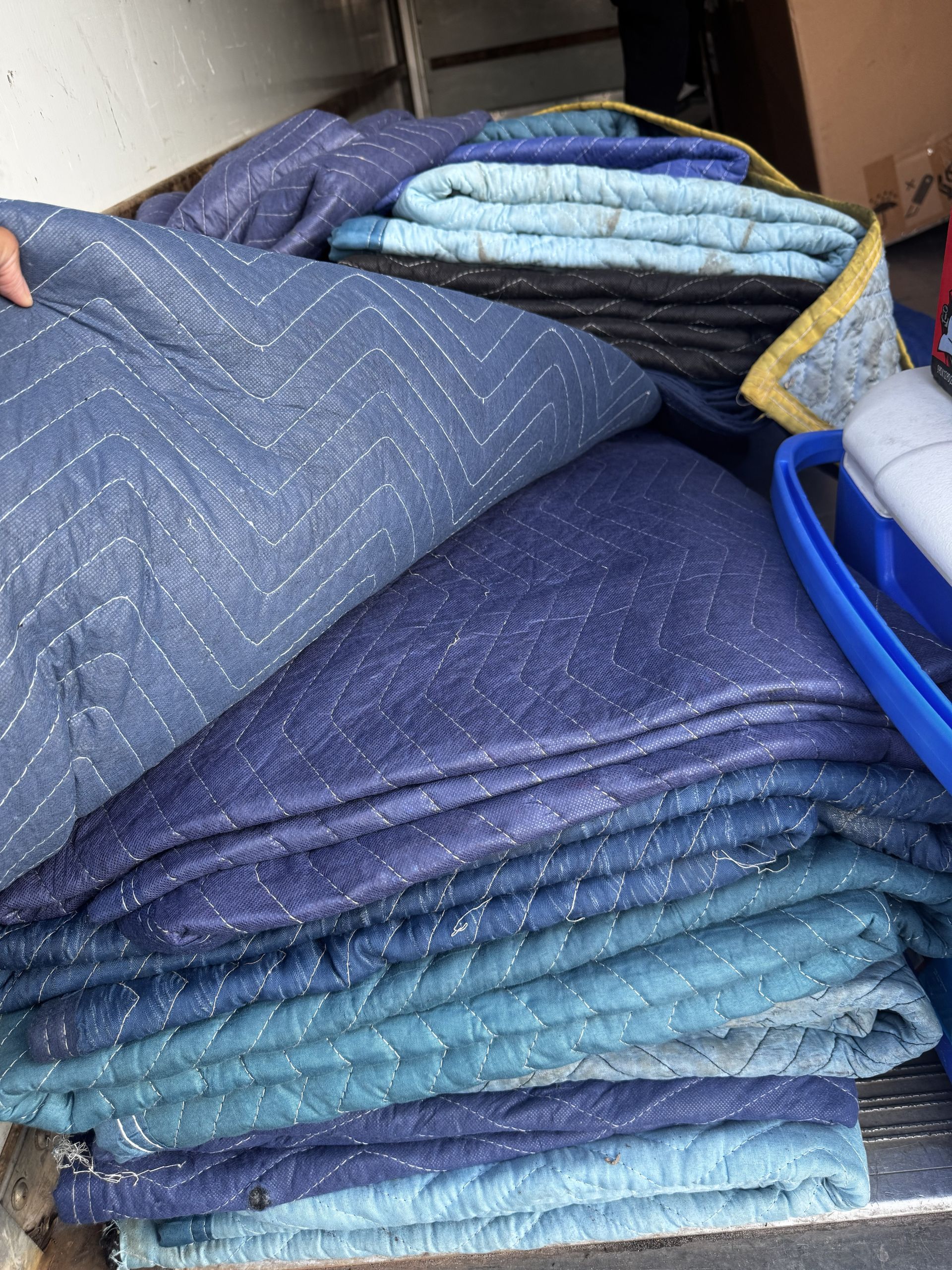 A stack of blue and teal quilted moving blankets, with a hand visible holding the corner of the top blanket.