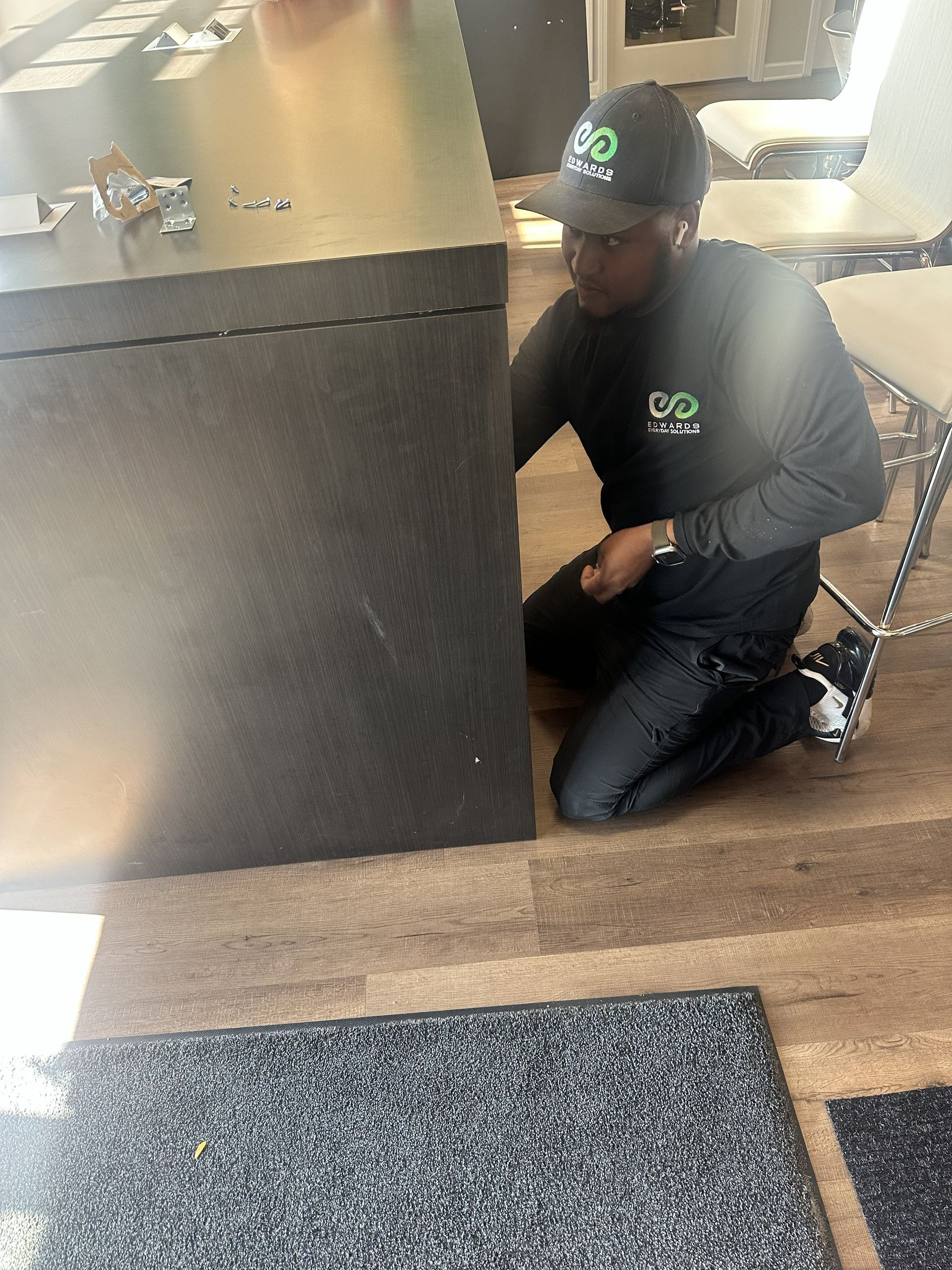 A person in a logo-branded gray cap and shirt kneels on a wooden floor, working at the base of a dark counter.