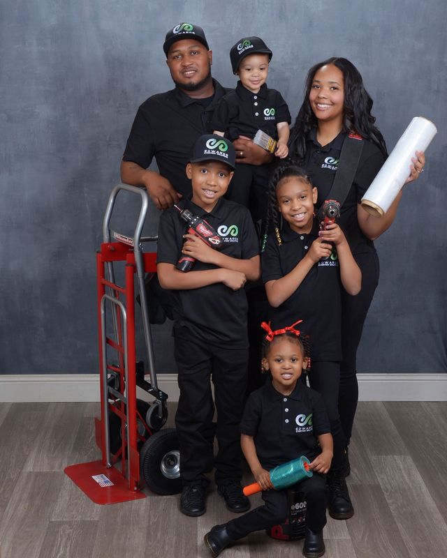 Family of six posing in matching black outfits beside a red hand truck against a gray backdrop