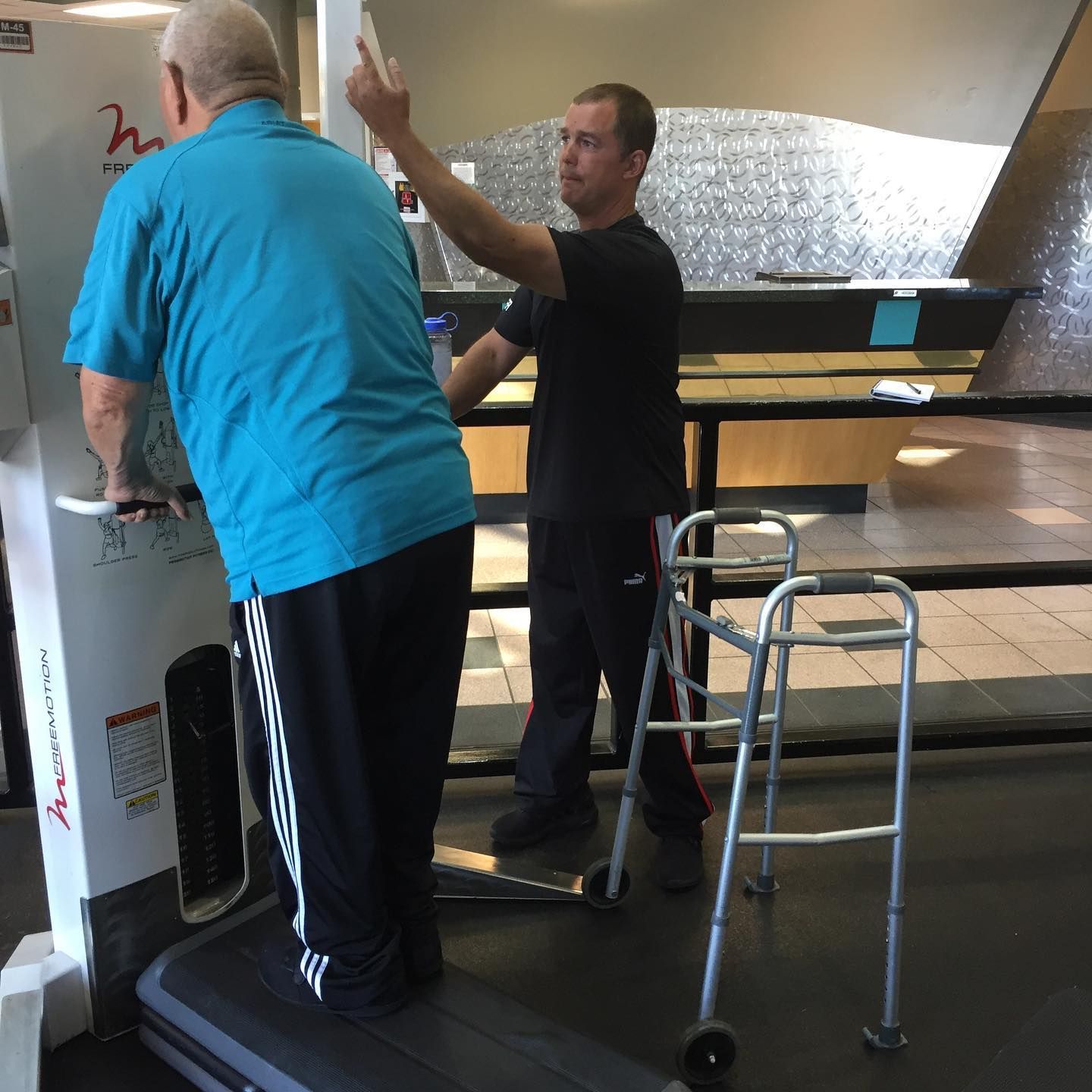 Two people using a standing desk near a walker in a bright room