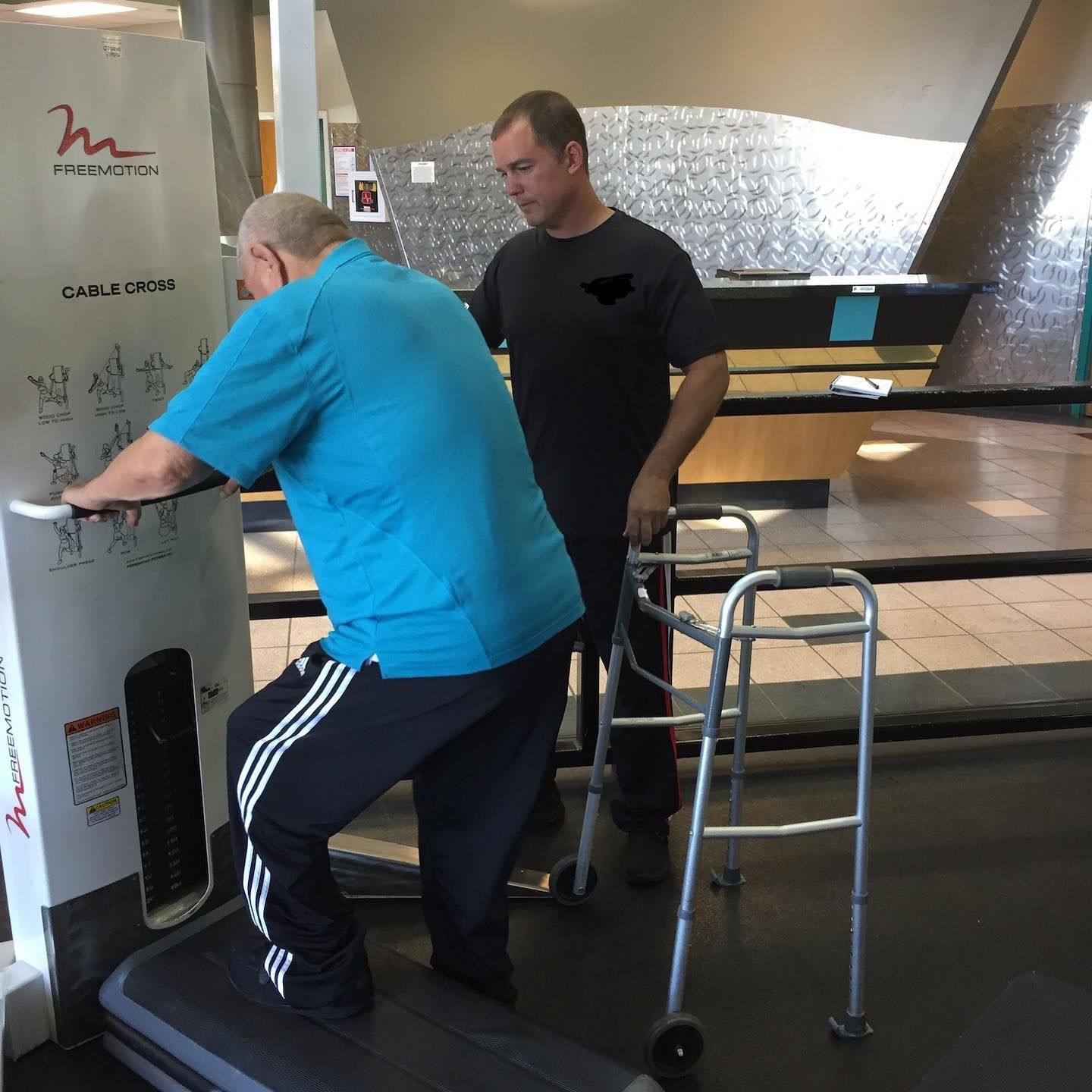 Two men using a gym machine, one in a blue shirt leaning on the equipment and one standing nearby with a walker.