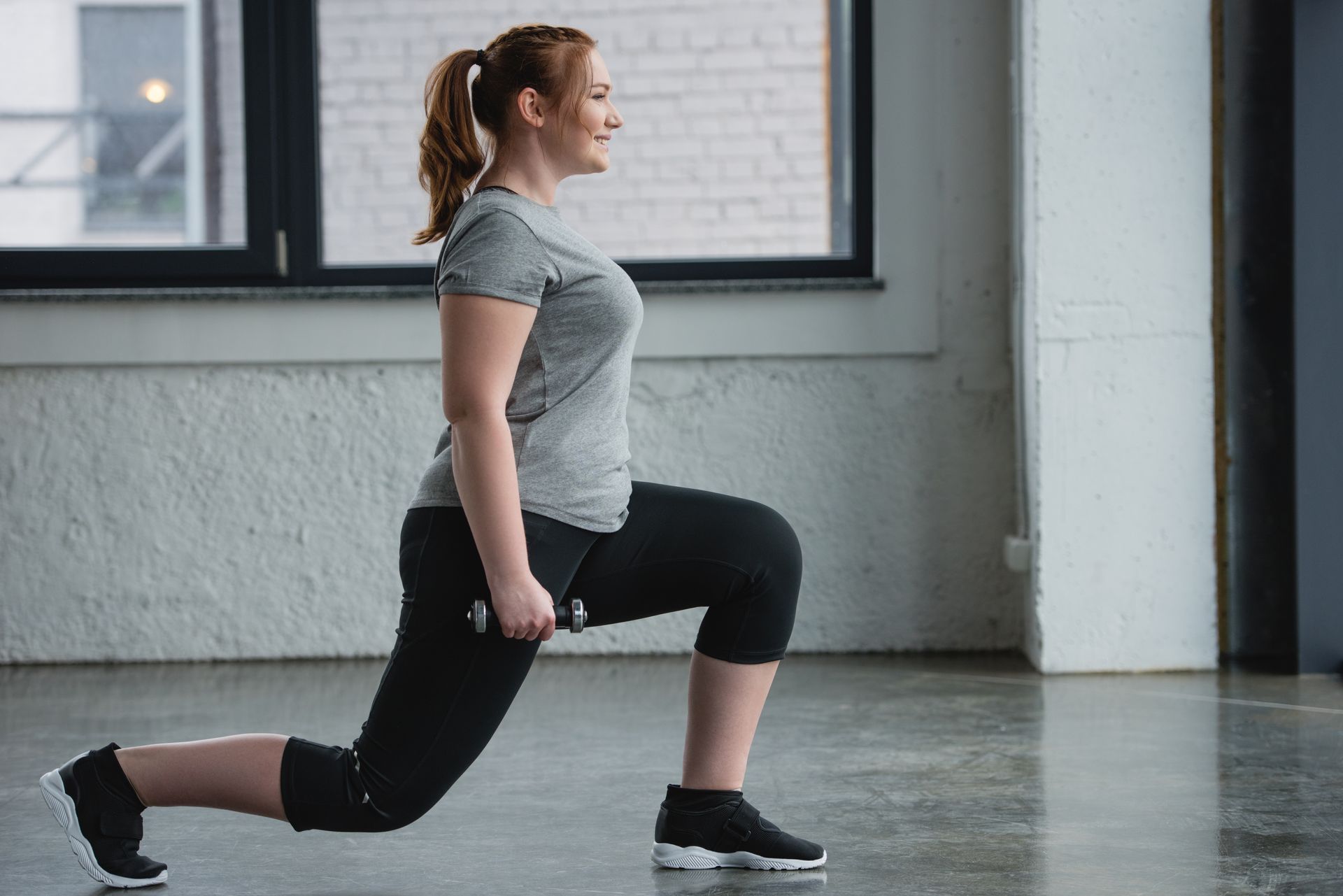 Person doing a forward lunge in a gym, wearing gray shirt and black leggings.