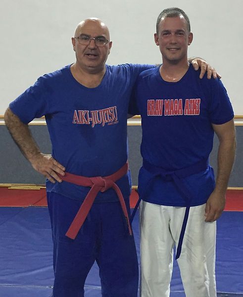Two martial artists in blue shirts posing on mats, one with a red belt and one with a blue belt.