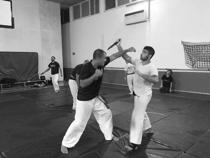 Two martial artists spar on blue mats in a gym, one blocking a raised stick strike.